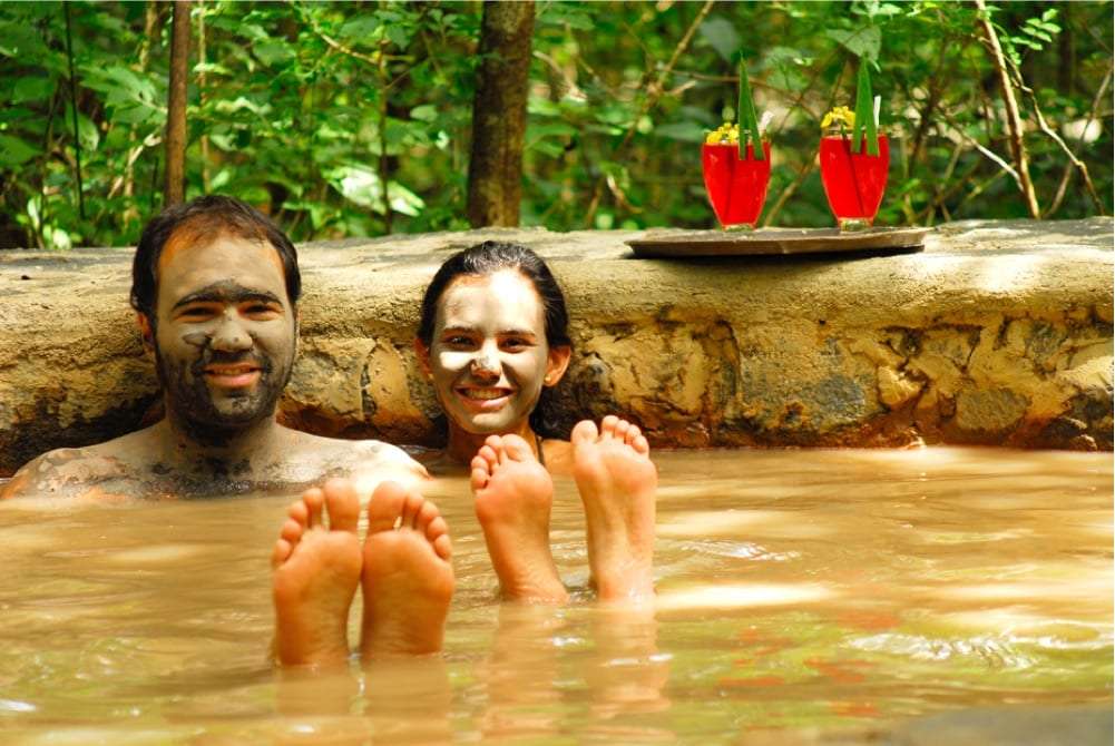 Couple with mud on their faces soaking in a warm pool with two red drinks behind them