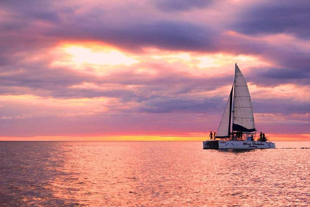 Catamaran with passengers sailing on calm water under a pink and purple sunset sky