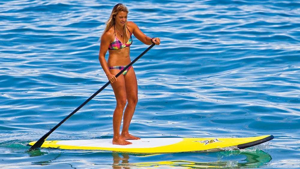 Woman in a bikini paddling a yellow stand up paddle board on blue water