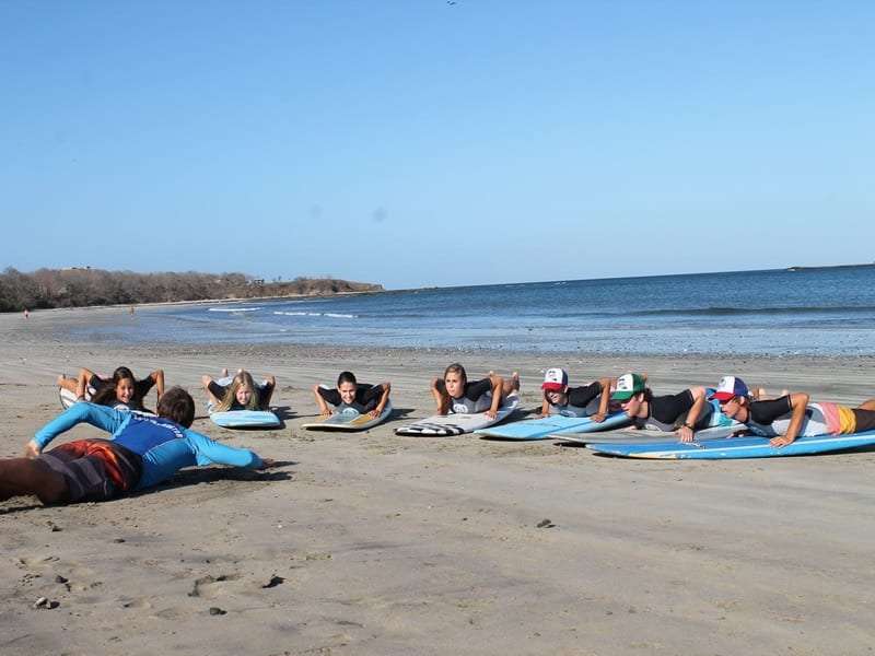 Surf students lying on boards on the sand practicing paddling while an instructor demonstrates