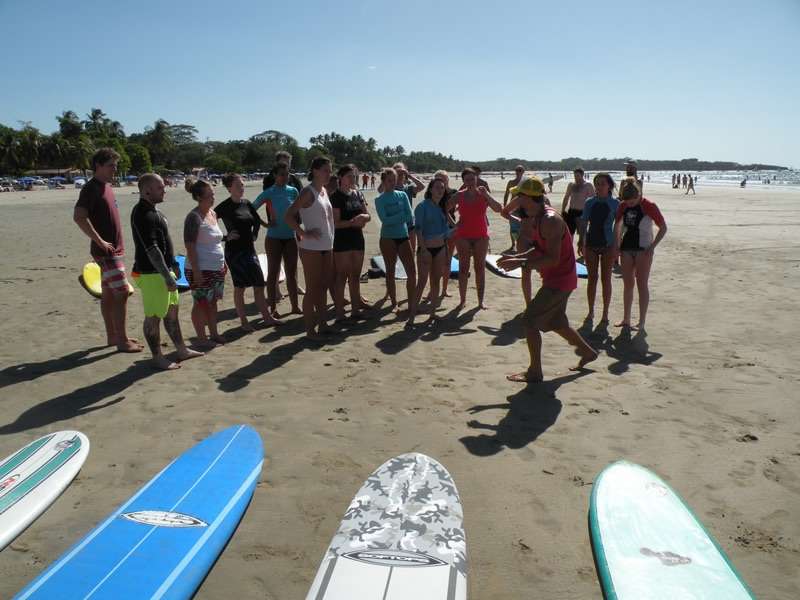Beach surf lesson with a coach addressing a large group of students in rash guards