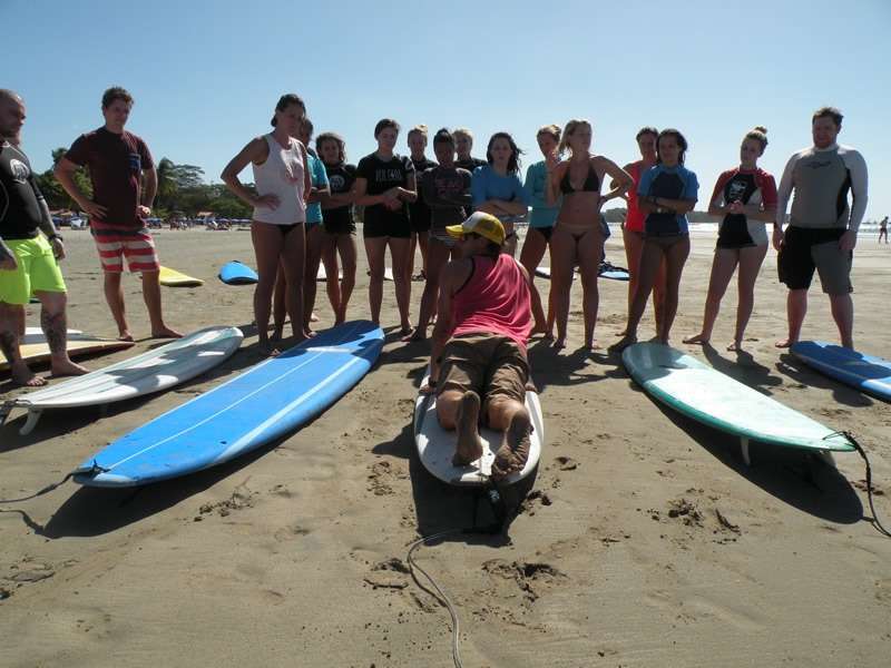Instructor lying on a surfboard demonstrating technique to students standing around him on the sand