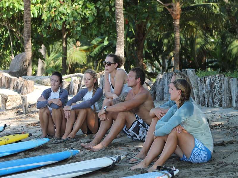 Surf students seated on the sand near palm trees listening to their instructor