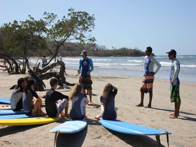 Three instructors and a group of students talking on a beach beside blue surfboards and driftwood