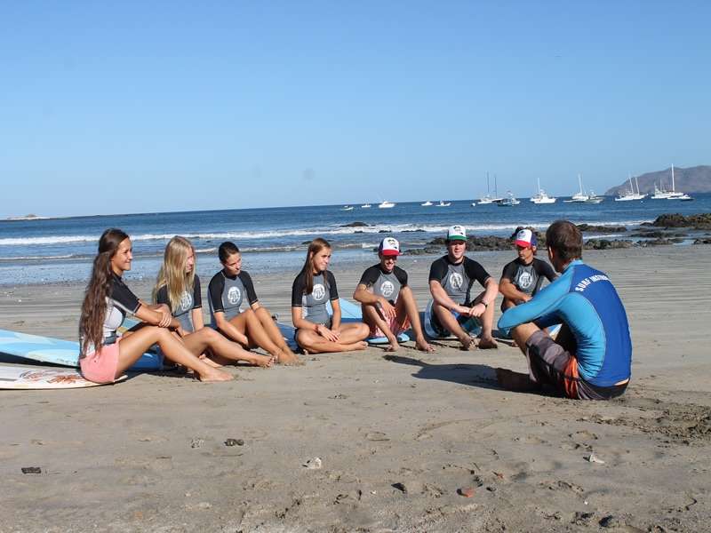 Students in matching rash guards sitting on the sand listening to an instructor, boats anchored behind