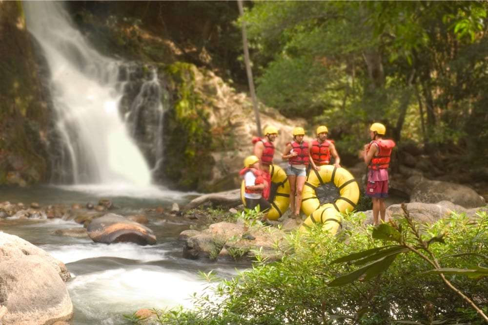 Tubing guests in life vests and yellow helmets beside yellow inner tubes at the foot of a waterfall