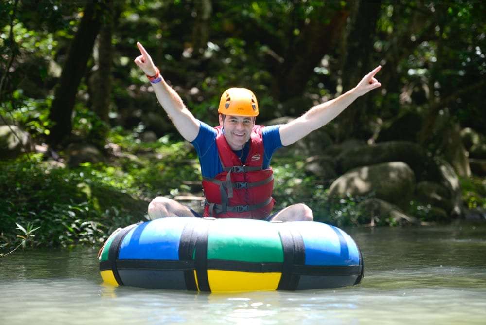 Smiling man in an orange helmet and life vest raising both arms while sitting in a colorful tube