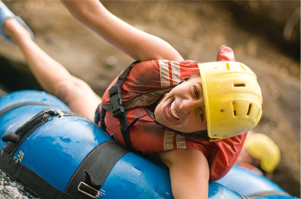 Woman in a yellow helmet and red life vest grinning as she rides a blue river tube