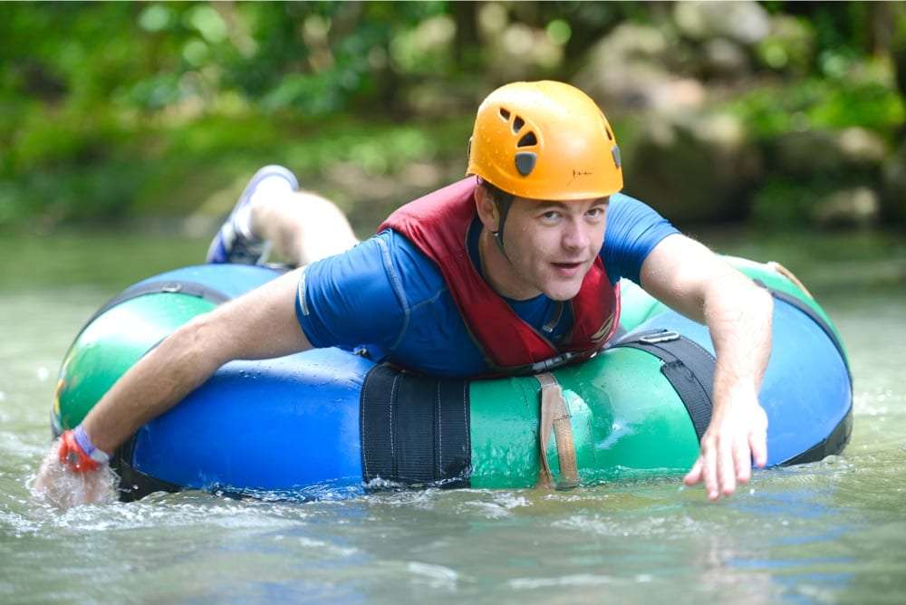 Man in an orange helmet and blue shirt paddling a green and blue tube on a shallow river