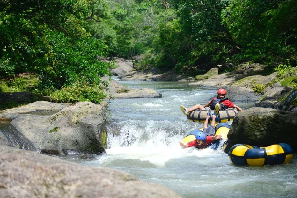 Two tubers in helmets riding rapids between large boulders on a forest river