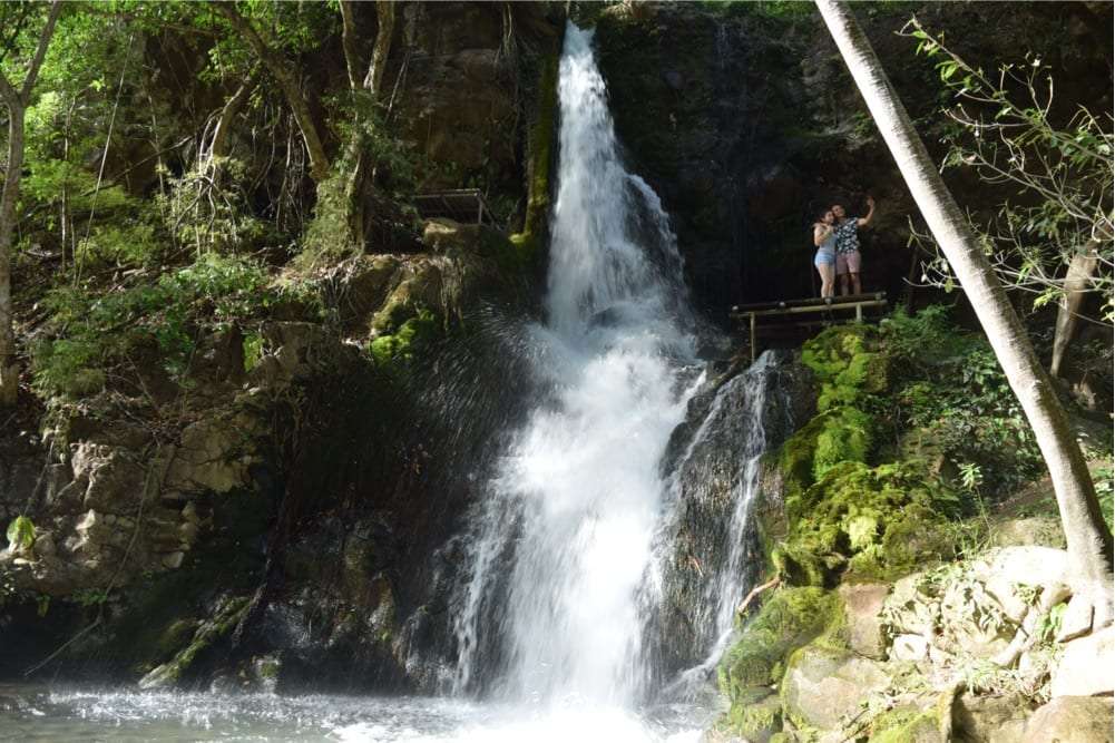 Two visitors on a wooden lookout beside a tall waterfall in the forest