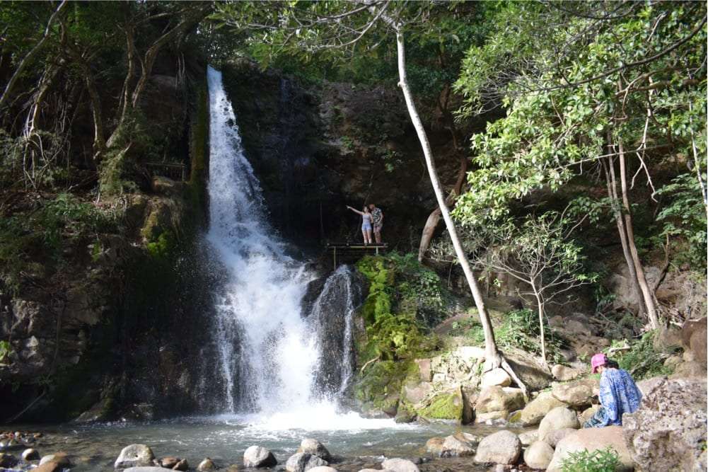 Visitors on a ledge above a waterfall with a person sitting by the rocky pool
