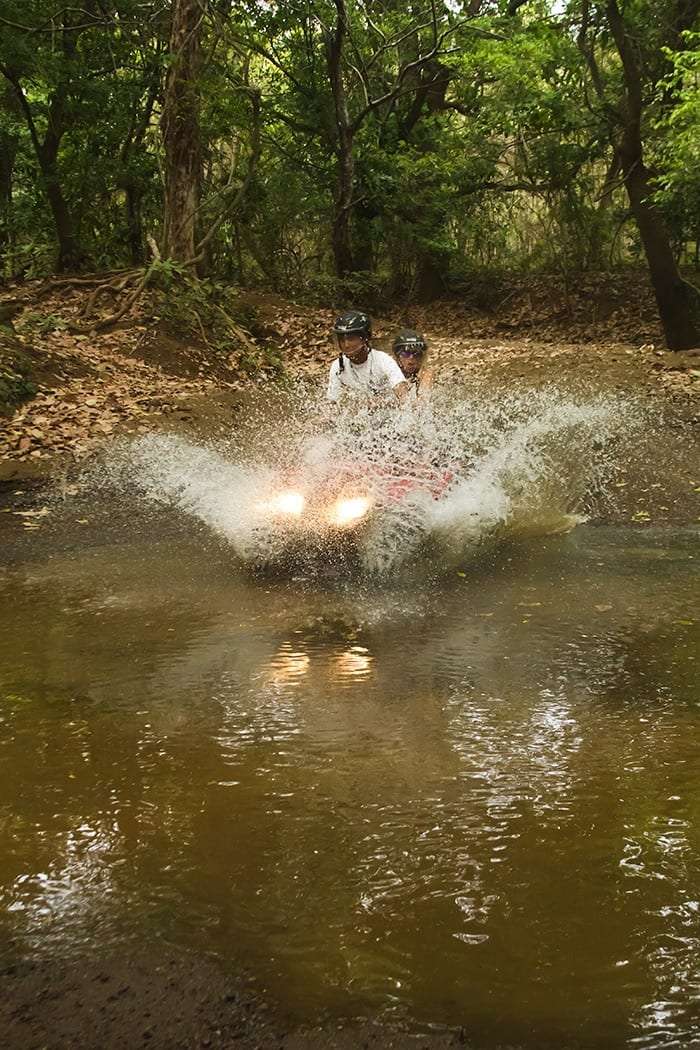 Red ATV splashing through a muddy puddle on a forest track with two riders