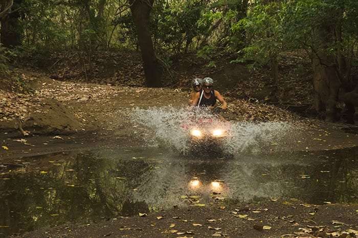 Two riders on a red ATV driving through a water-filled dip in a wooded trail