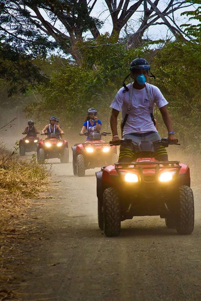 Guide on a red ATV leading a line of riders along a dusty road under trees