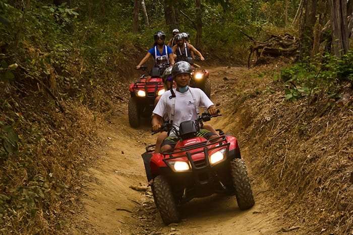 Riders on red ATVs descending a narrow, sunken dirt trail between dry banks