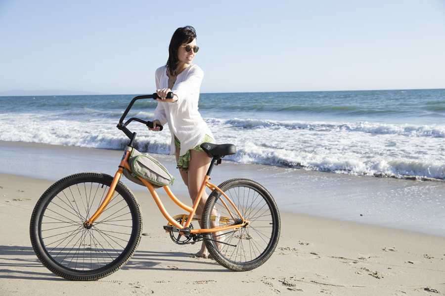 Woman in sunglasses standing beside an orange beach cruiser bicycle on the sand by the sea