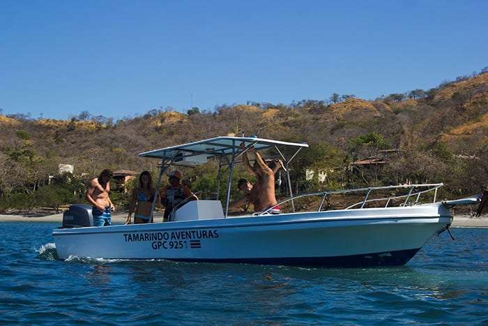 Passengers standing and posing on a white boat near a dry hillside and sandy beach