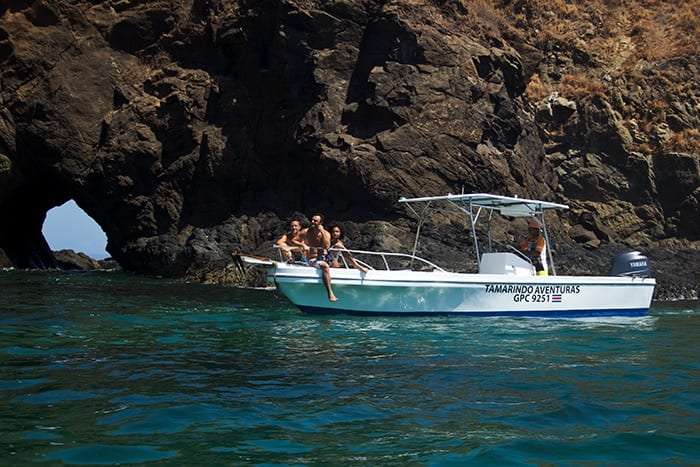 White boat with passengers beside a dark cliff with a sea arch in turquoise water