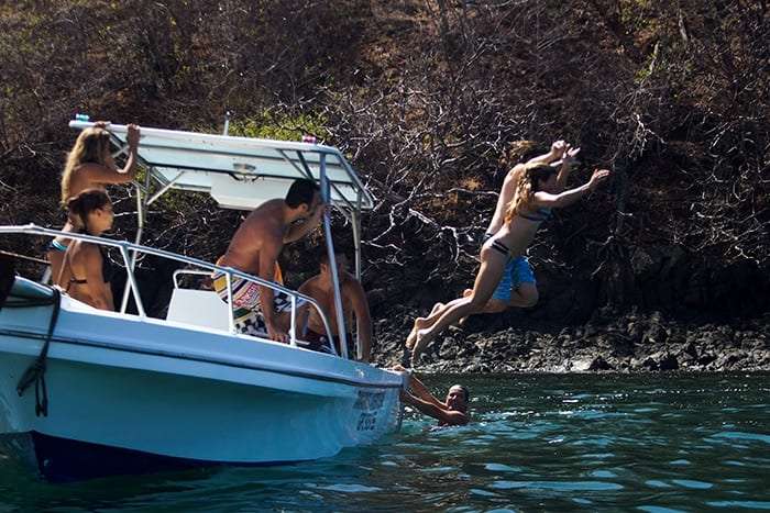 Woman leaping off a boat toward a rocky shore while a swimmer waits in the water