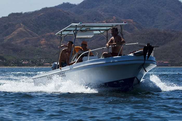 Boat carrying passengers cutting through waves with a hillside coast behind