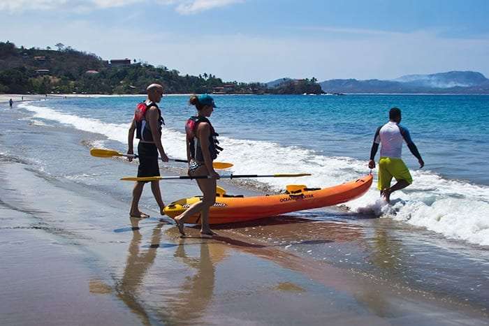 Kayak guests and a guide in a yellow shirt walking an orange sea kayak into the surf