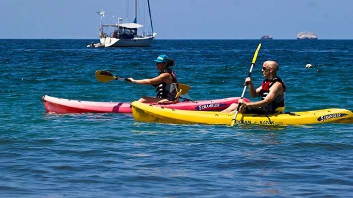 Two people paddling a pink and a yellow kayak on blue water with a sailboat behind