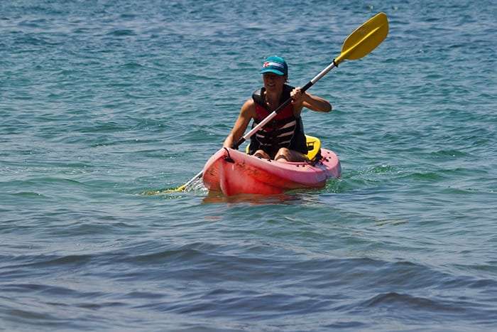 Smiling kayaker in a teal cap and life vest paddling a pink kayak on calm water