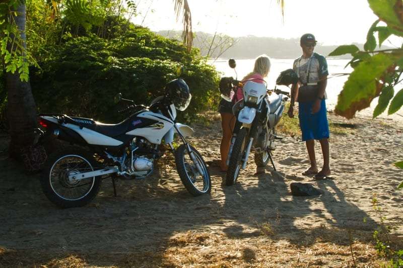 Two dirt motorbikes parked on a sandy beach with a woman and a man in sandals talking