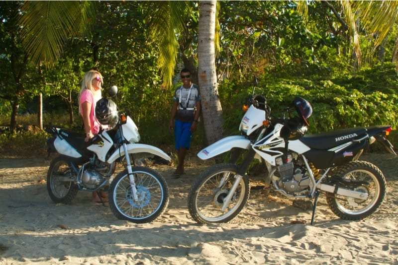 Woman and man standing beside two white Honda motorbikes under palm trees on sand