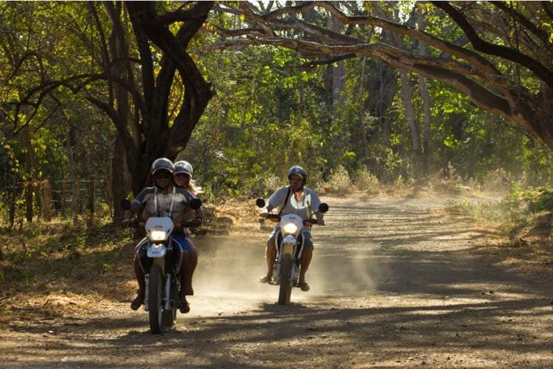 Two motorbikes with riders on a dusty dirt road under overhanging trees