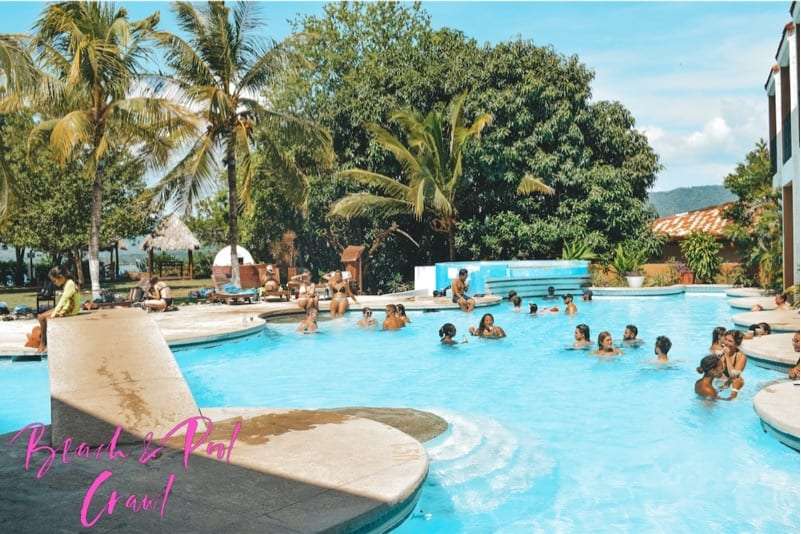People swimming and lounging in a large blue pool with palm trees around it