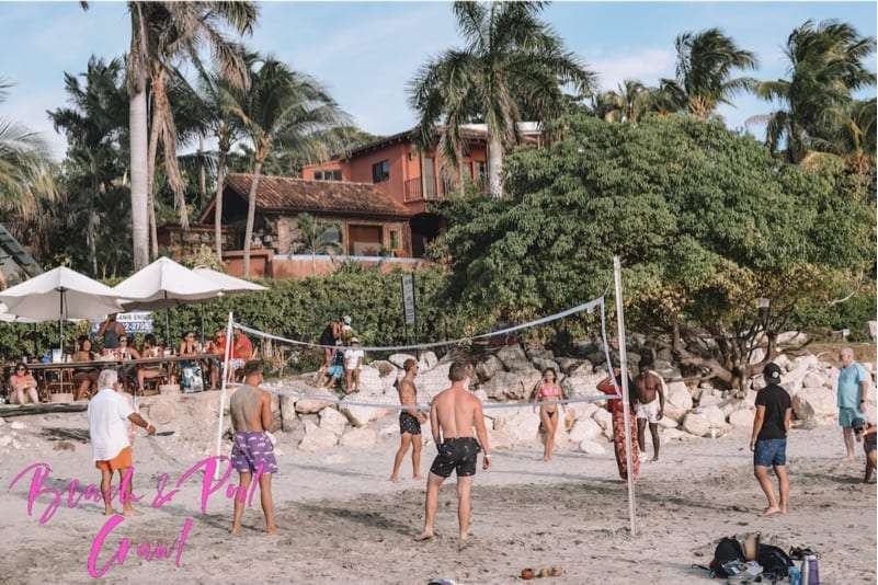 Beachgoers playing volleyball on the sand with palm trees and a house behind