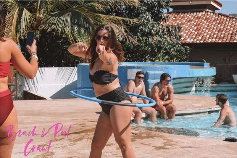 Woman in a black bikini spinning a hula hoop beside a pool as others relax behind her