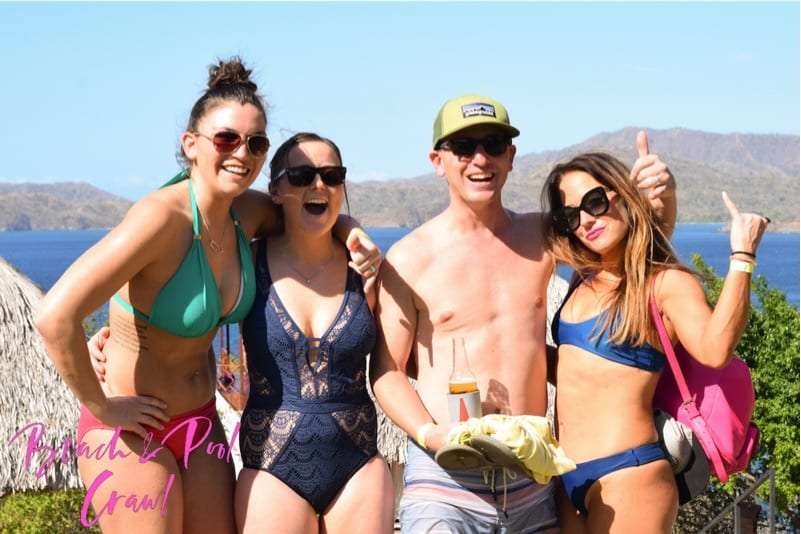 Three women and a man in swimwear smiling with a bay and hills behind them