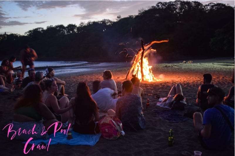 Group sitting on the sand around a bonfire at dusk beside a dark forest