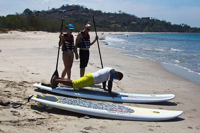 Instructor doing a push-up on a paddleboard while two students stand on the beach holding paddles