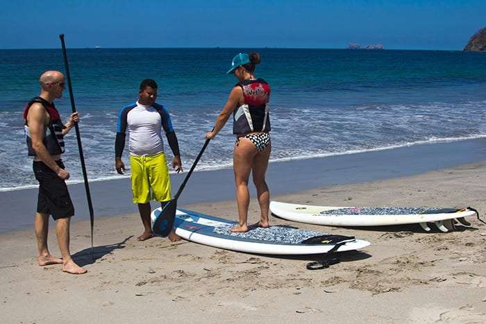 Man and woman in life vests with paddles listening to an instructor beside paddleboards on the beach