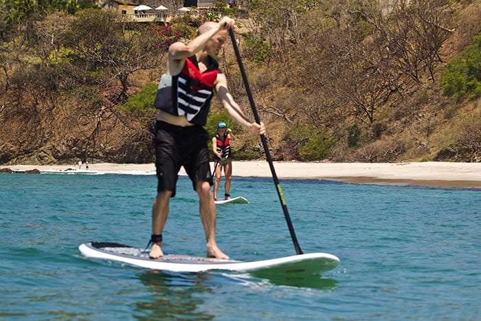 Man in a life vest standing on a paddleboard with a second paddler behind him near a sandy cove