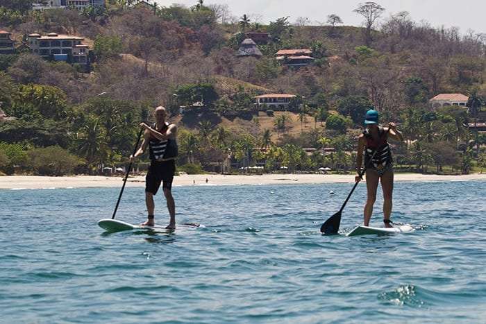 Man and woman standing on paddleboards on calm water in front of a hillside with houses