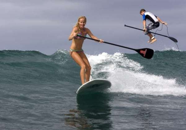 Woman standing on a paddle board riding a small wave, with a child falling off behind