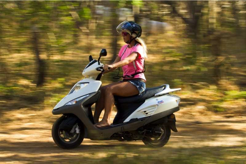 Woman in a pink top and helmet riding a white scooter along a blurred forest road