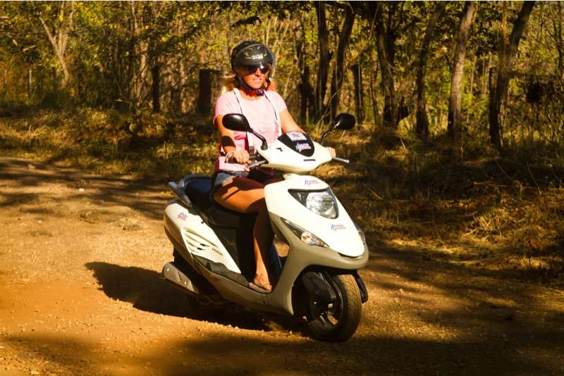 Woman in a helmet and sunglasses riding a white scooter on an orange dirt road
