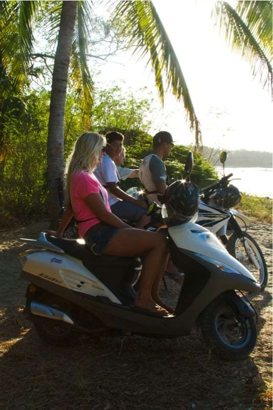 Woman on a scooter beside two men and a motorbike under a palm tree at the shore