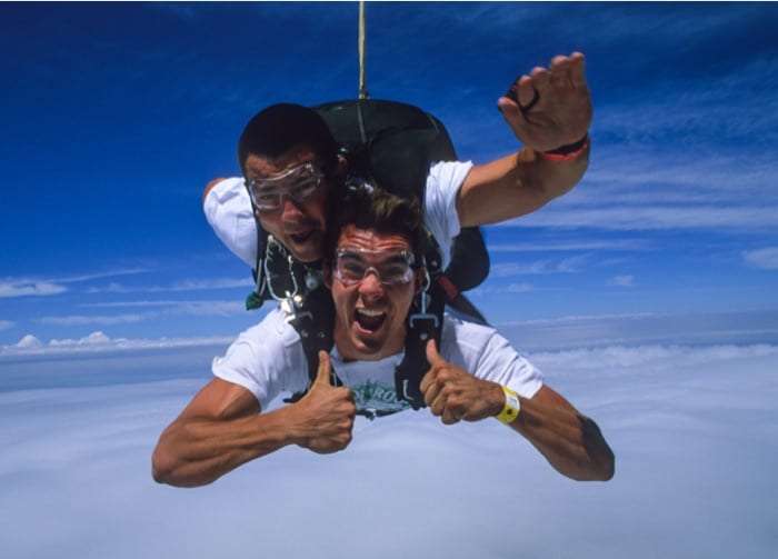 Tandem skydivers giving thumbs up in freefall above a layer of clouds