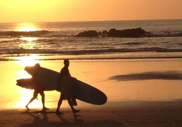 Two people carrying surfboards along a beach at sunset