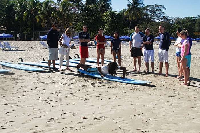 Group of students watching an instructor lie on a surfboard on the sand to demonstrate technique