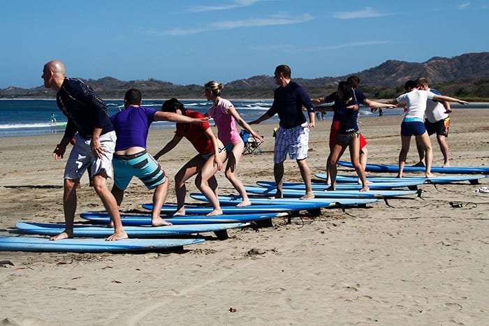 Students practicing standing up on surfboards on the sand while an instructor guides them