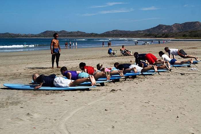 Row of students lying on surfboards on the beach while an instructor looks on