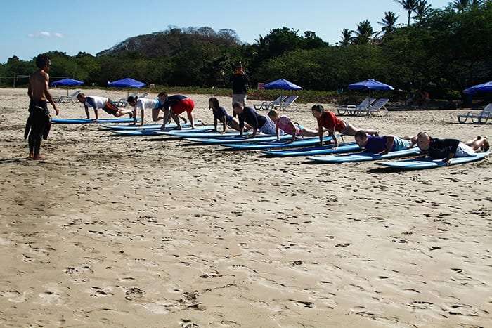 Students doing push-up positions on surfboards on the sand beside blue beach umbrellas